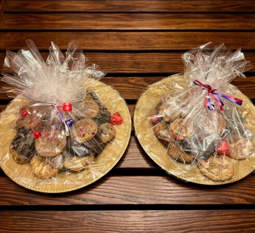 Two gold trays of homemade cookies with heart sprinkles and foil-wrapped chocolate hearts, covered in plastic wrap on a wooden table.